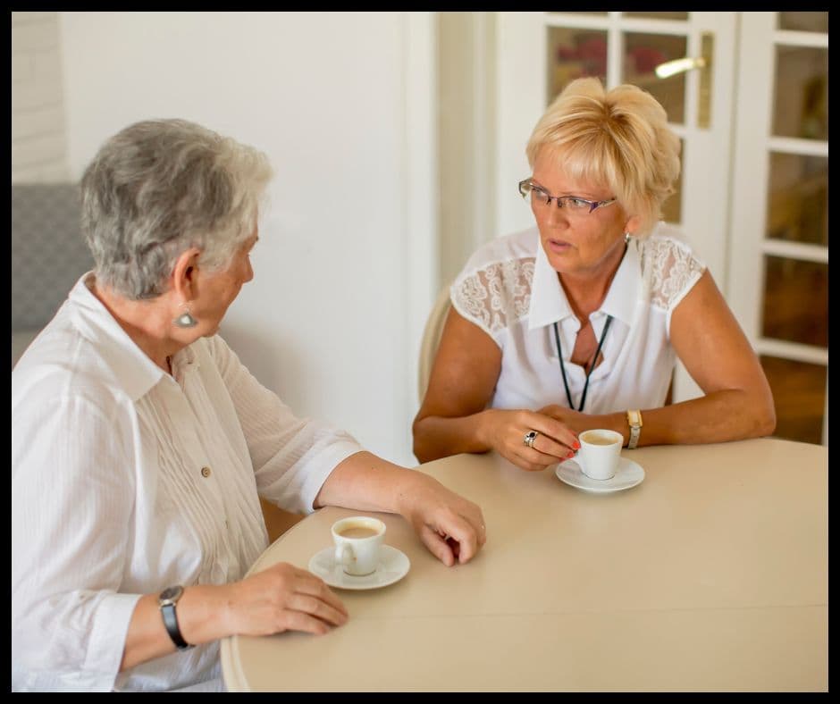 Two senior women sit at a table, drinking coffee and engaging in a serious conversation.
